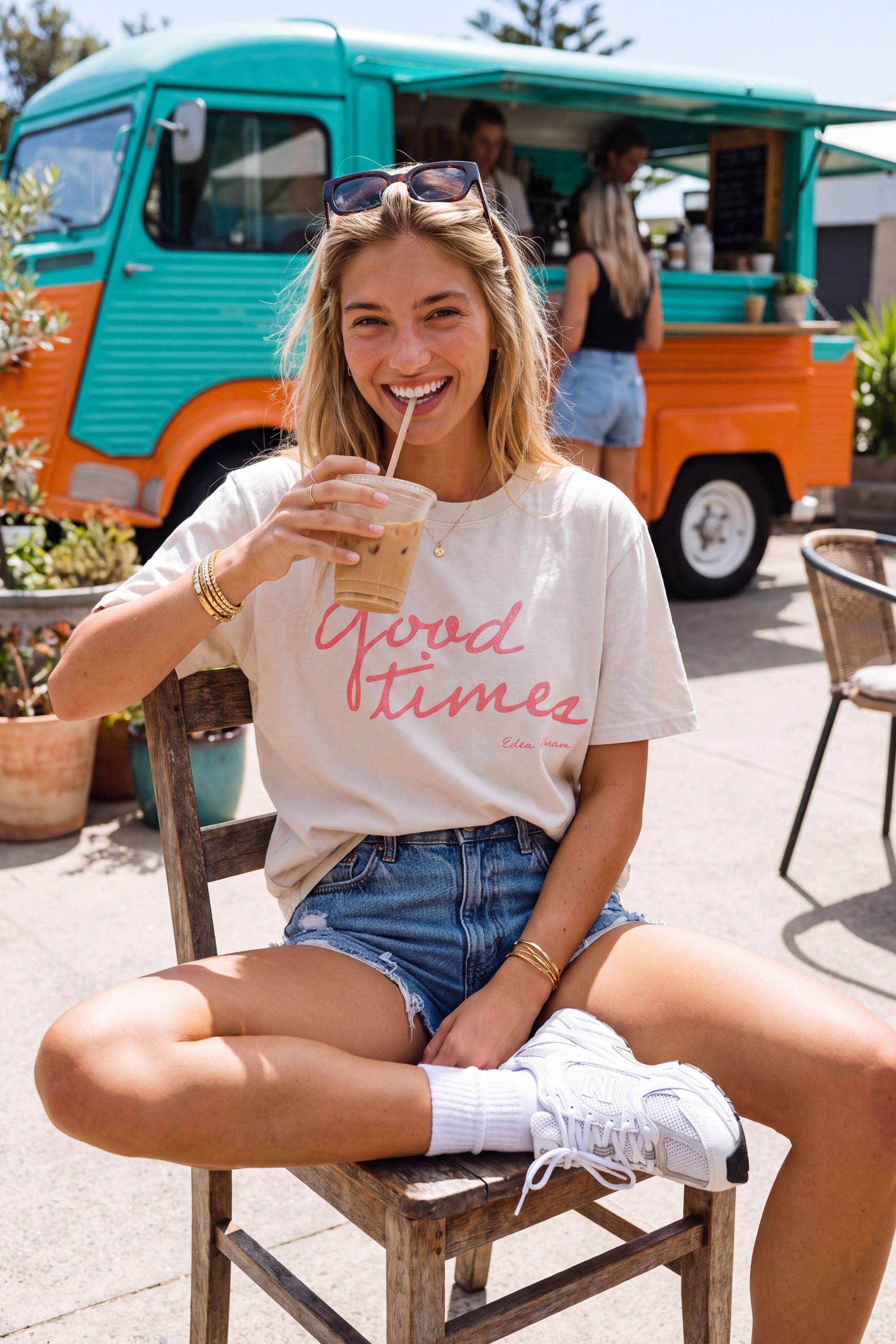 A woman sitting on a wooden chair in front of a turquoise and orange food truck, wearing a cream graphic t-shirt with pink script text that reads 'Good times' and denim shorts.