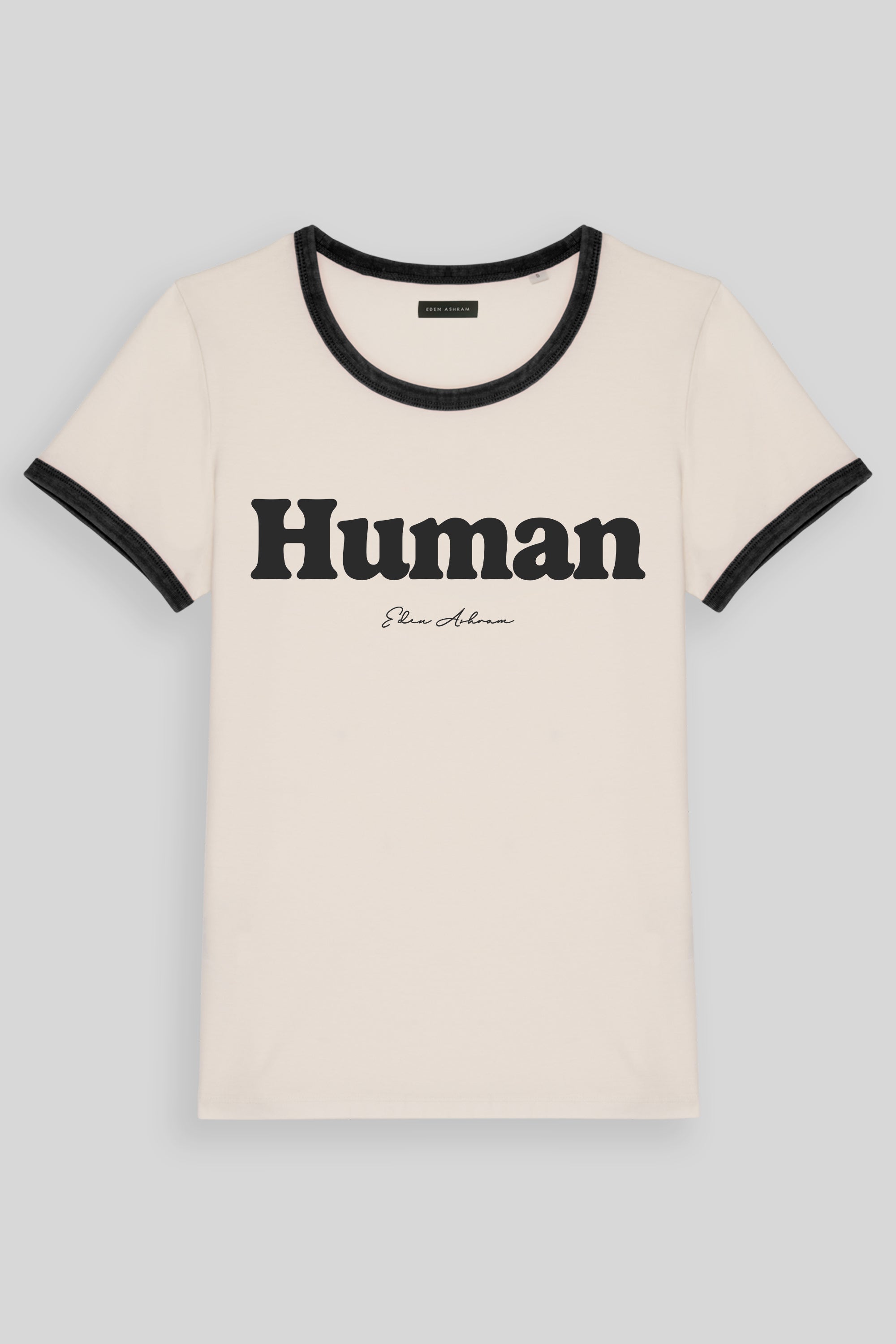 An off-white ringer t-shirt with black trim on the crew neck and sleeve cuffs, featuring the word 'Human' printed in bold black letters across the chest, with a small cursive Eden Ashram brand name underneath.